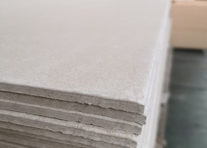 Stack of thick gray drywall or board sheets stacked on a pallet in a workshop or warehouse setting, viewed from the side.